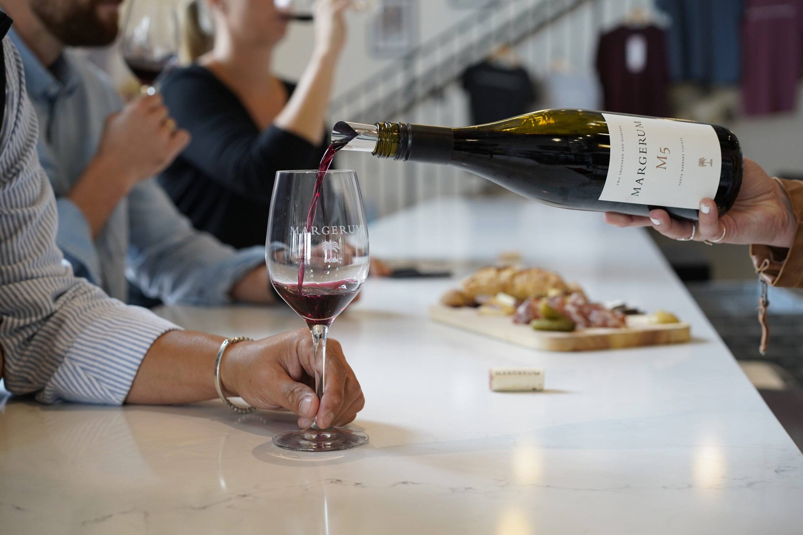a hand with a bracelet is holding the stem of a wine glass while red wine is being poured into the glass. in the blurred background is a cheese and charcutier board and a woman drinking red wine from a wine glass. the background is the margerum tasting room.