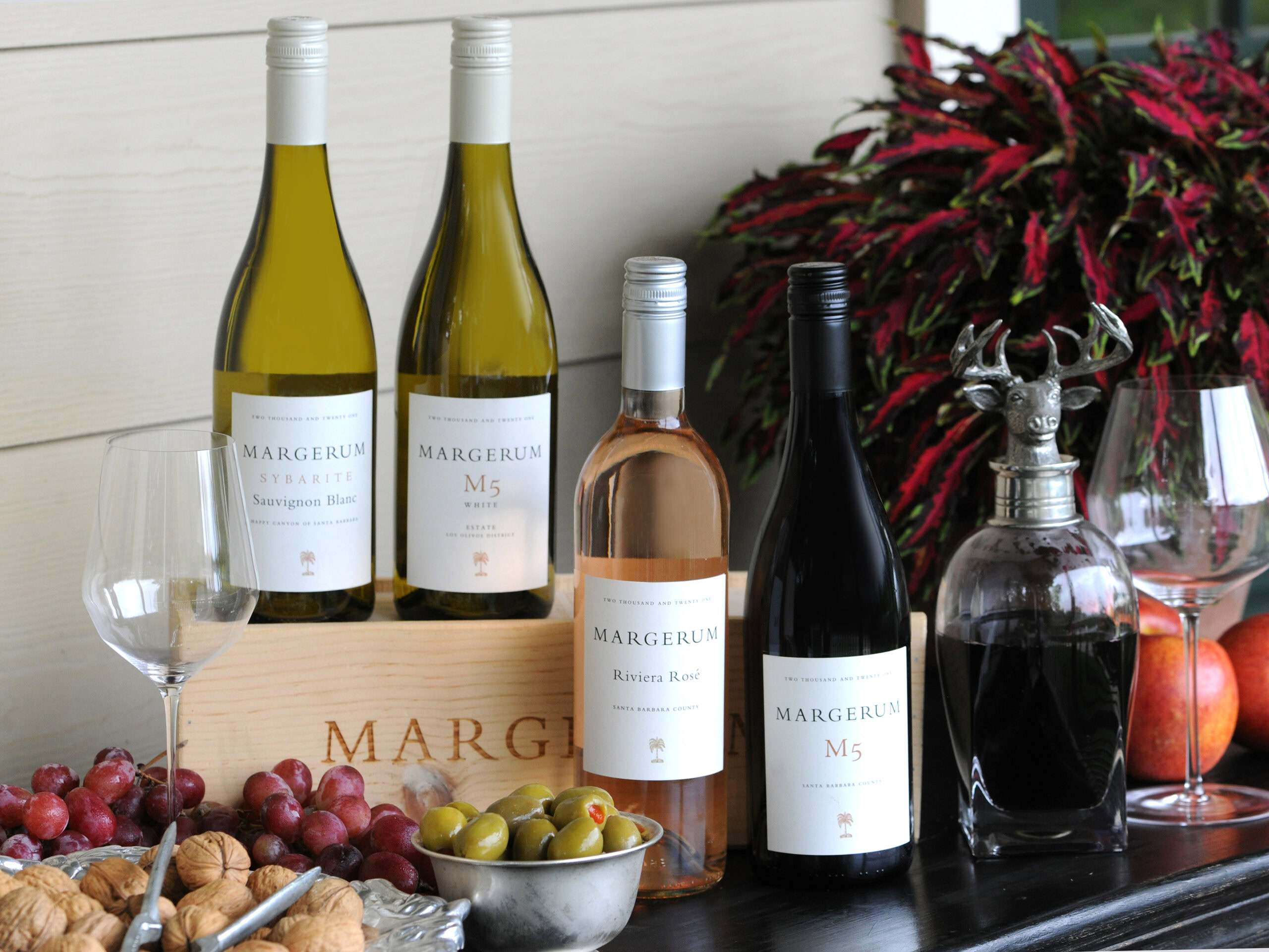 four bottles of margerum wines, two white wines, one rose, and one red wine with a red plant behind them and olives, grapes and crackers in front.