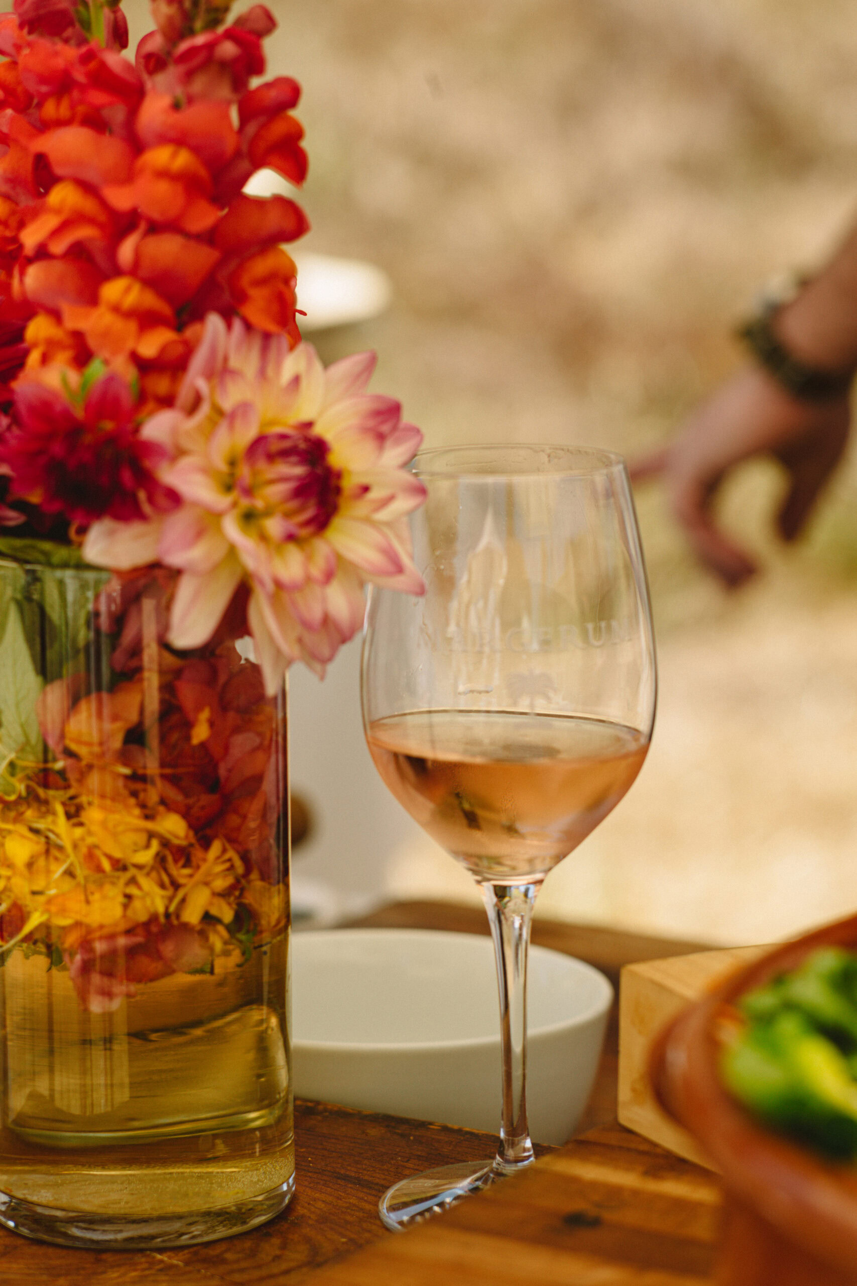 a pour of rose in a wine glass with pink and orage flowers next to it, the background is blurred.