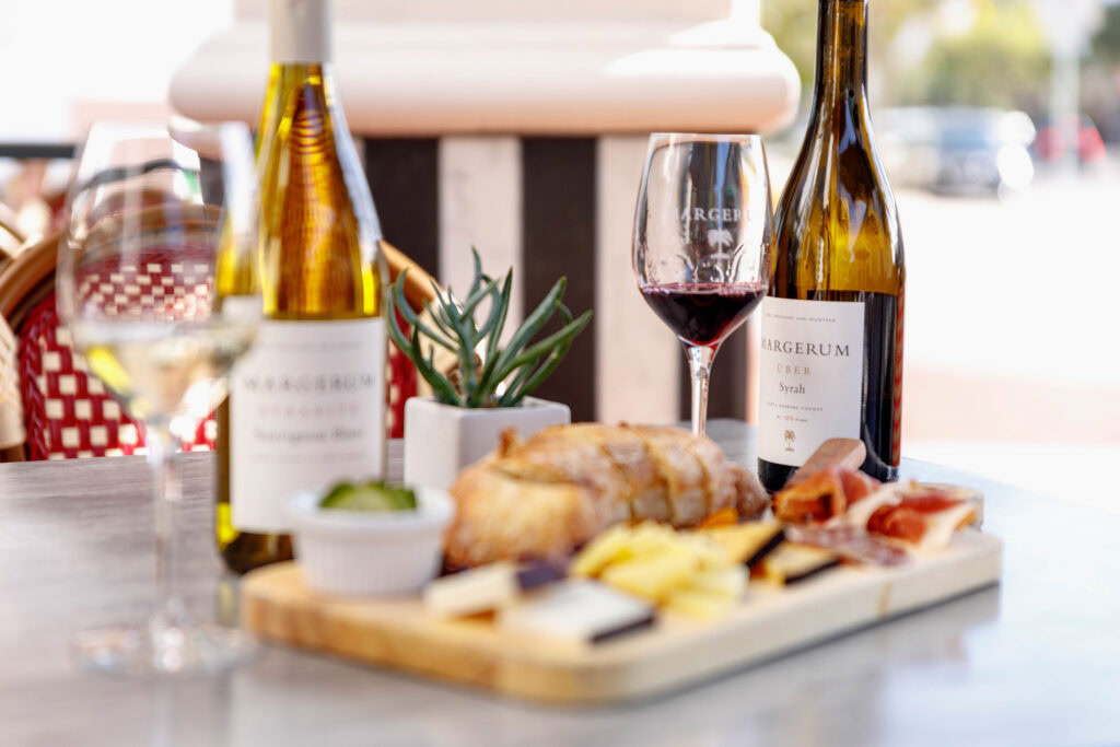 two margerum bottles, one white and one red with two classes of wine, in front of it is a cheese and charcuterie board
