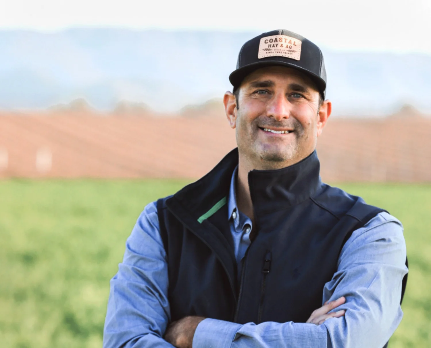 Man in blue vest, wearing a hat with his arms crossed in a vineyard