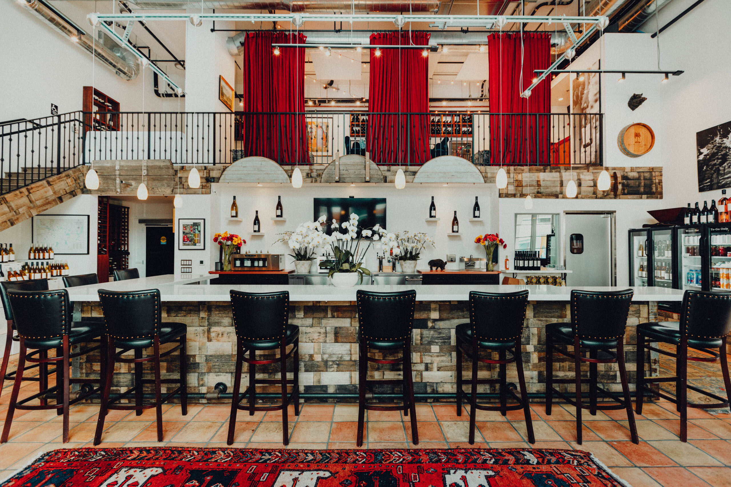 Margerum Tasting Room Bar - there are seven dark barstools visible, on top of a red rug that is draped over light tan flooring. The bar is white and above the bar is the upstairs mezzanine that has red curtains draping.